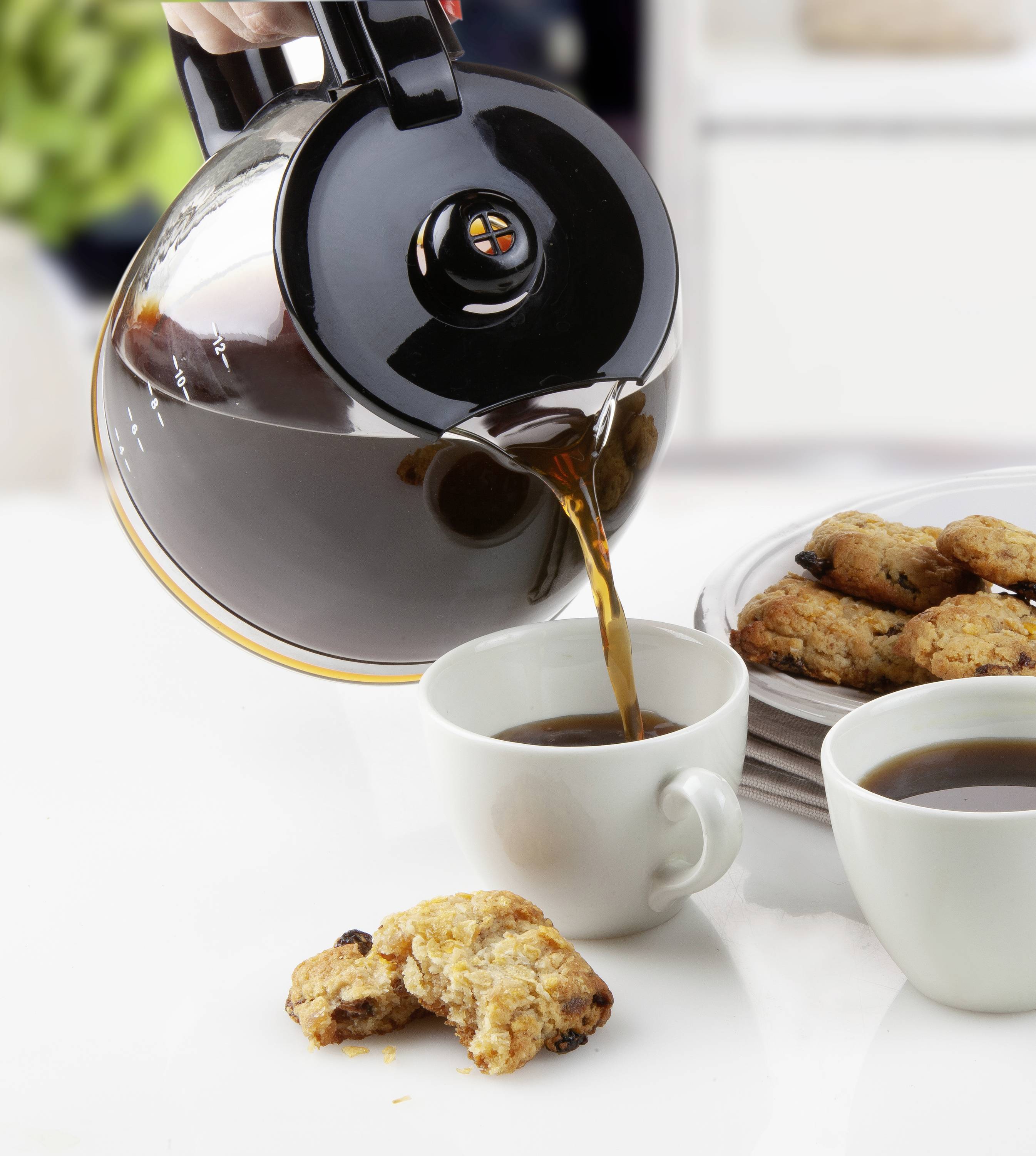 Ruka nalévá kávu z konvice do šálku. Vedle stojí talíř s cookies. Světle osvětěné, útulné kuchyňské prostředí.