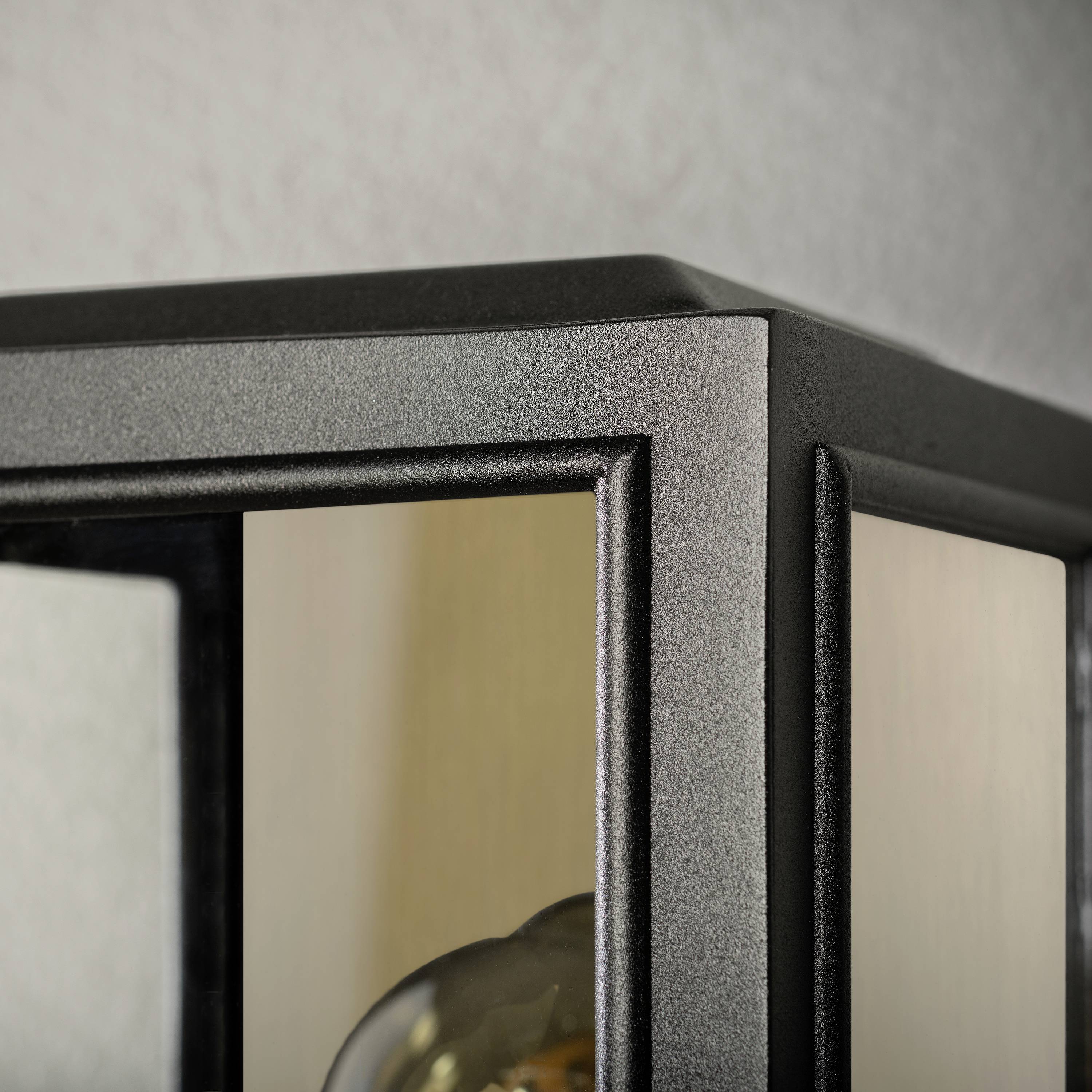 Detailní záběr černé hranaté lampy se skleněnými výplněmi. Uvnitř je viditelná žárovka.