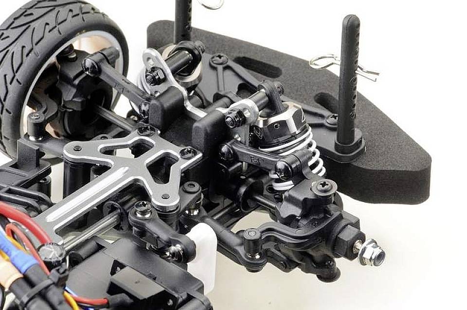 Detail záběr přední nápravy dálkově ovládaného automobilu, zobrazující tlumiče, zavěšení a kolo s viditelnými komponenty a kabeláží.