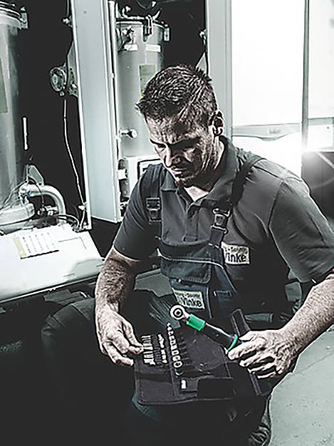 Muž v pracovních montérkách držící nástroj, soustředěný a pracující na strojním zařízení, osvětlený v průmyslovém prostředí.