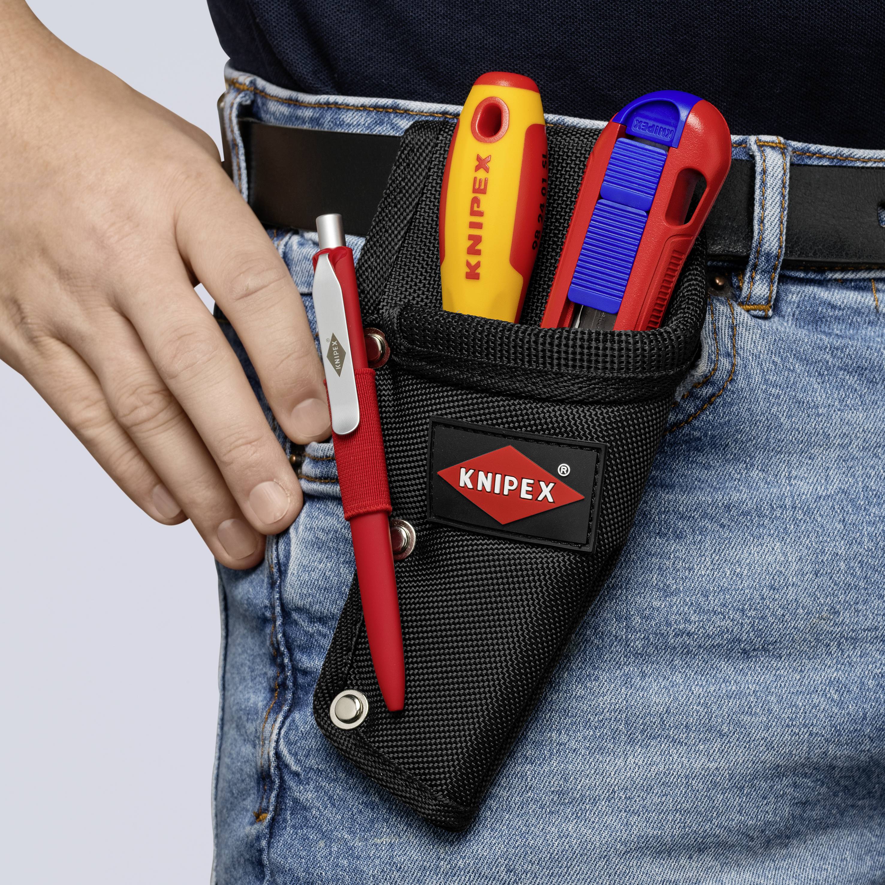 'Pouzdro na nářadí připevněné k pásku s KNIPEX skládacím nožem, řezacím nožem a perem, které nosí osoba v džínách a tmavém horním dílu oblečení.'