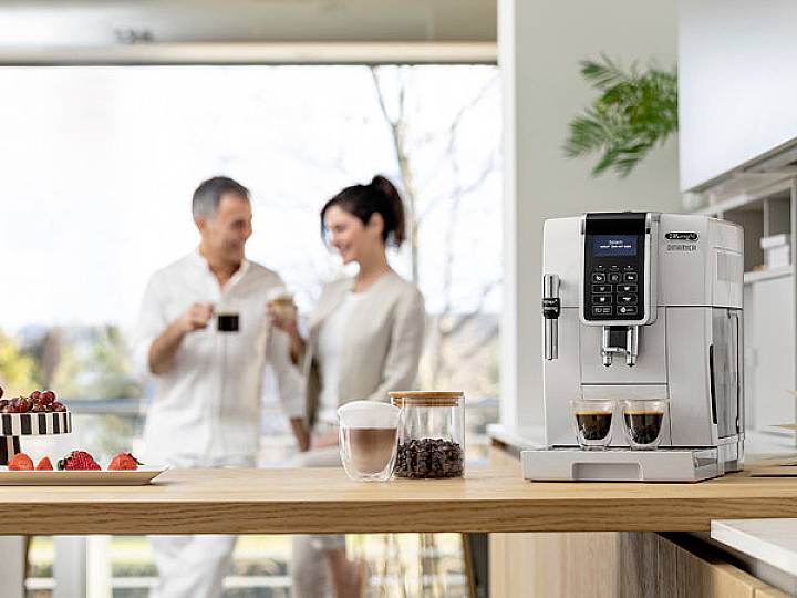 Pár popíjí kávu v moderní kuchyni. V popředí stojí kávovar připravující dvě šálky kávy.