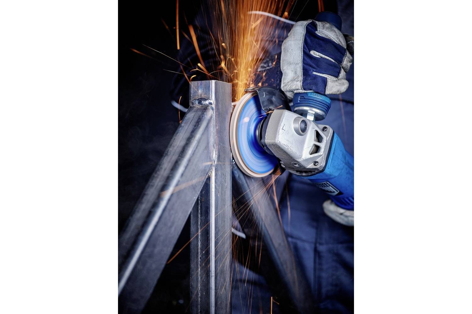 Jedna osoba brousí kov kotoučovou bruskou. Z brusného kotouče létají jiskry. Osoba má na sobě bezpečnostní rukavice.
