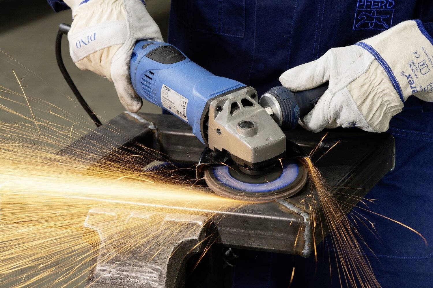 Pracovník v modré pracovní kombinéze brousí kov úhlovou bruskou. Kolem létají jiskry, pracovník je vybaven ochranným oděvem.