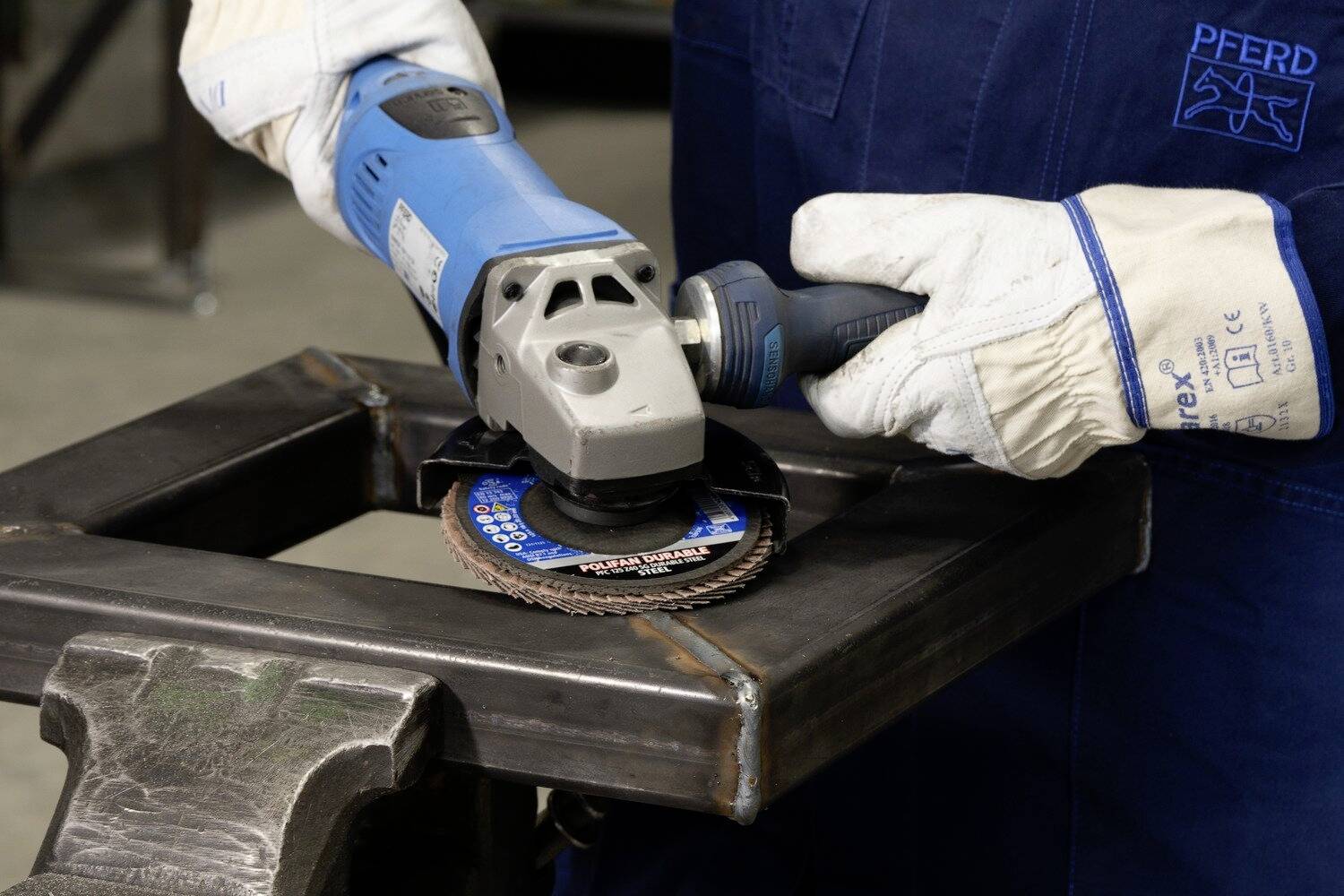 Osoba brousí kovovou rámovou strukturu úhlovou bruskou. Má na sobě ochranné rukavice a pracovní oděv.