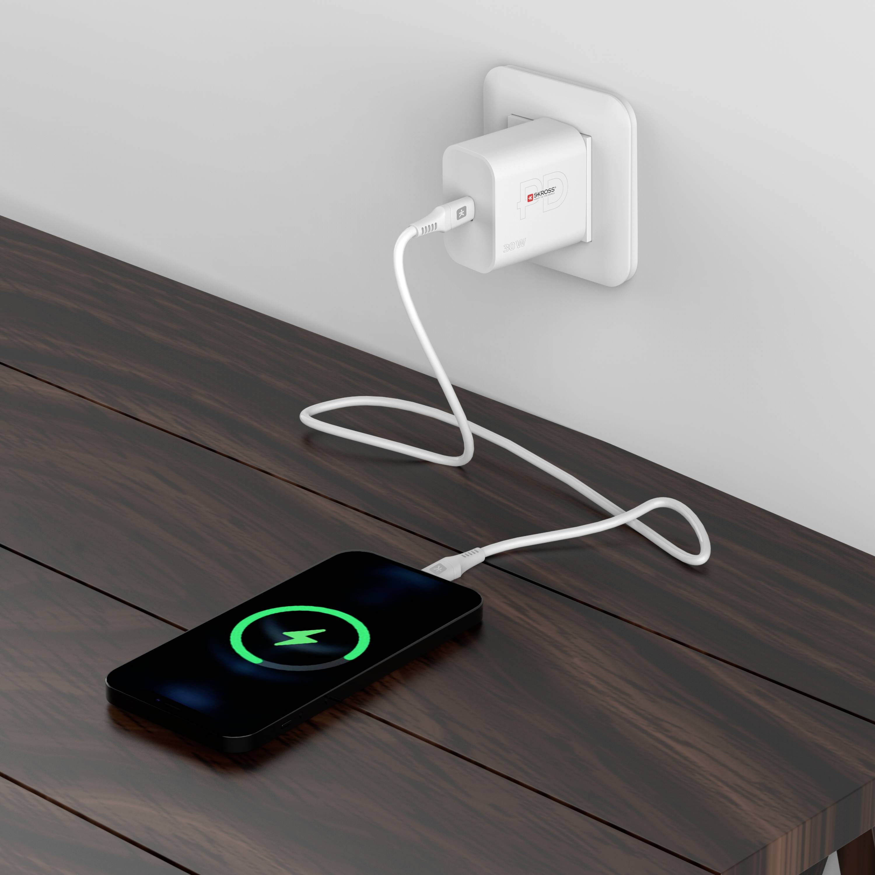 Mobilní telefon se nabíjí na dřevěném stole. Je připojen pomocí USB kabelu k adaptéru zapojenému do nástěnné elektrické zásuvky.