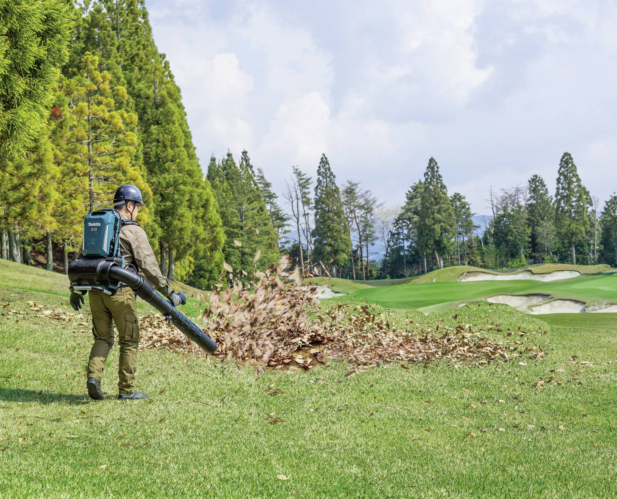 Osoba fouká listí z trávníku pomocí foukače. V pozadí jsou stromy a golfové hřiště.