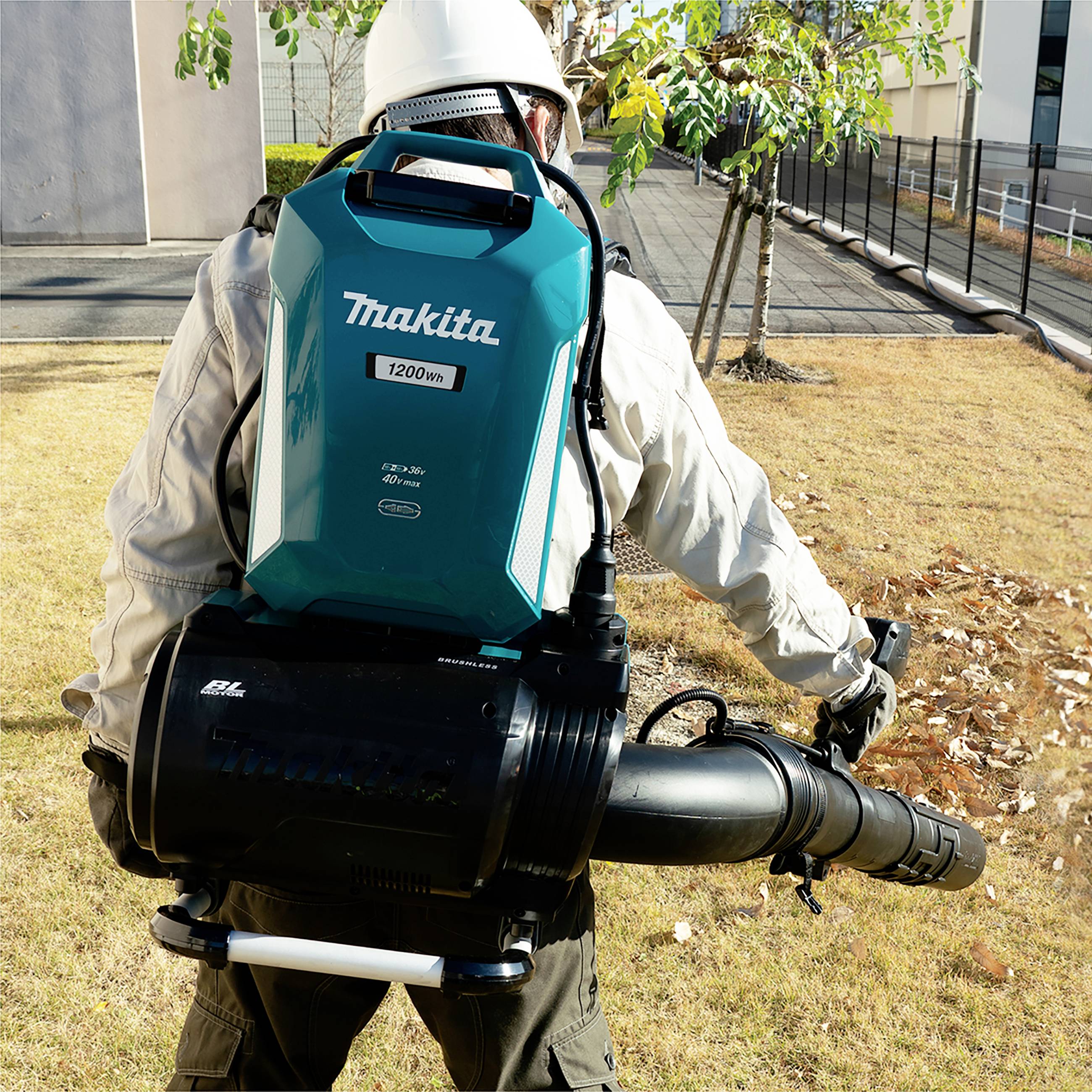 Muž fouká listí listovým fukarem Makita 1200W na trávníku. V pozadí je kancelářská budova s několika stromy.