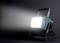 Přenosná pracovní lampa vyzařuje jasné světlo doleva. Lampa je modrá a černá, s pevnou rukojetí.