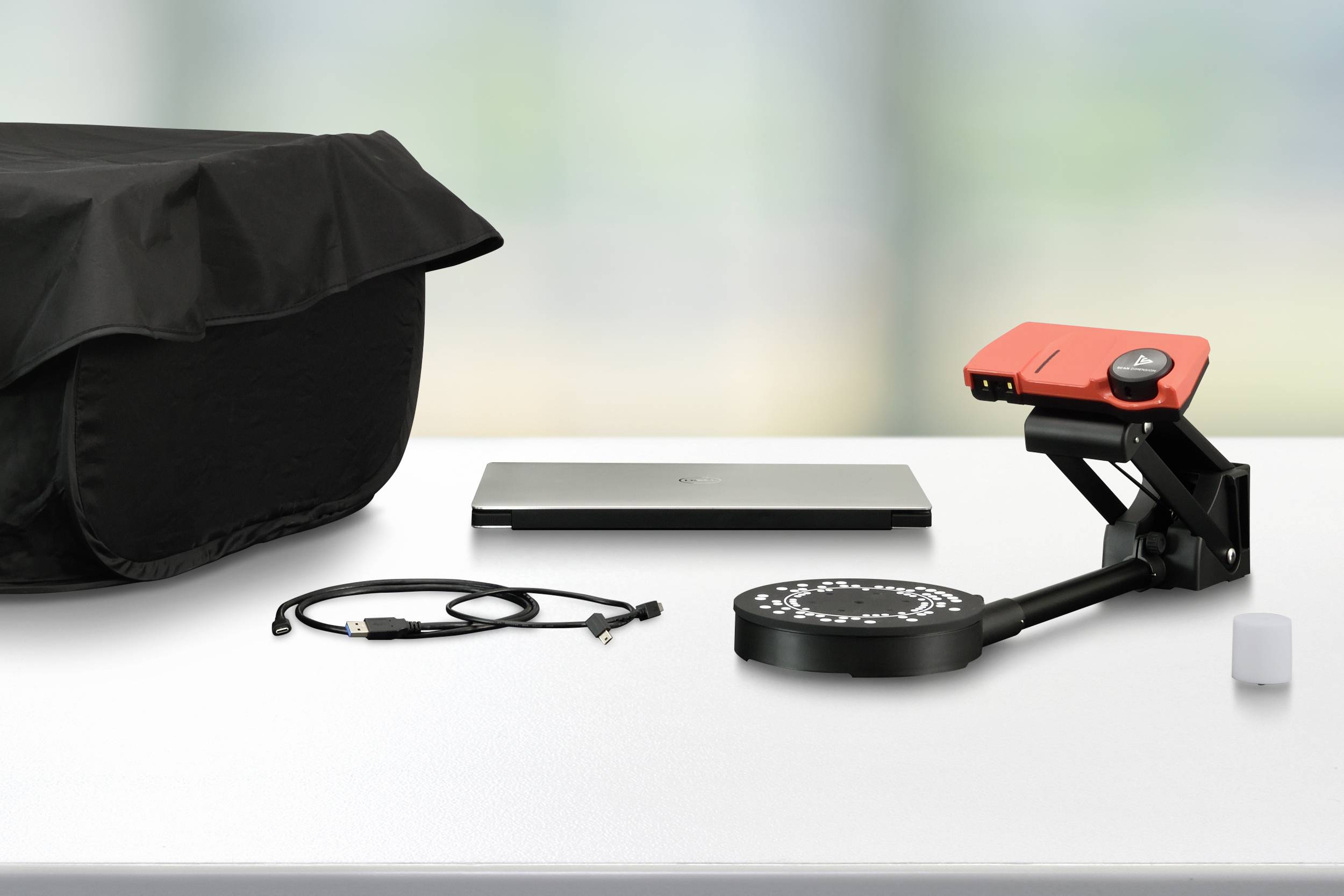 Skener s červeným pouzdrem, USB kabelem, krytem, laptopem a bílou kostkou na stole před rozmazaným pozadím.