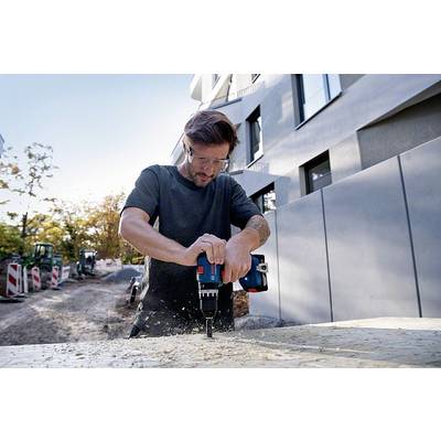 Muž vrtá elektrickou vrtačkou do dřevěné desky na staveništi. V pozadí jsou viditelné stavební přístroje.