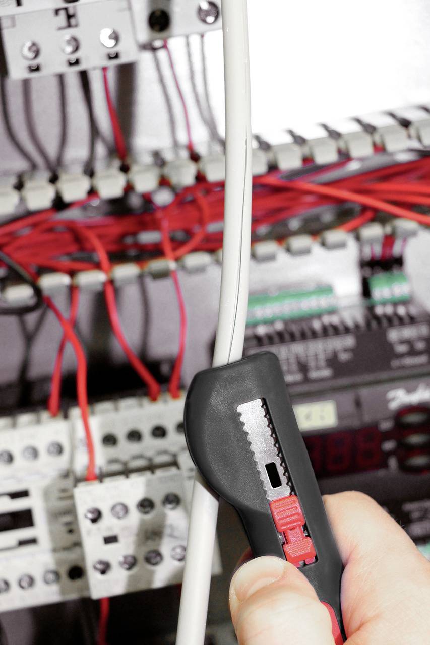 Osoba drží černý nůž a stříhá bílý kabel v otevřené rozvodné skříni s červeným a bílým elektrickým vedením.