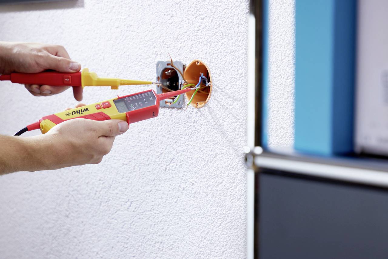 Osoba provádí měření elektrických vodičů v nástěnné zásuvce pomocí digitálního multimetru.