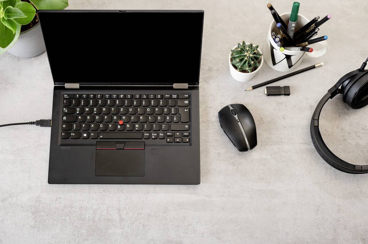 Laptop s černou obrazovkou stojí na psacím stole, obklopen rostlinami, sluchátky a kancelářskými potřebami.