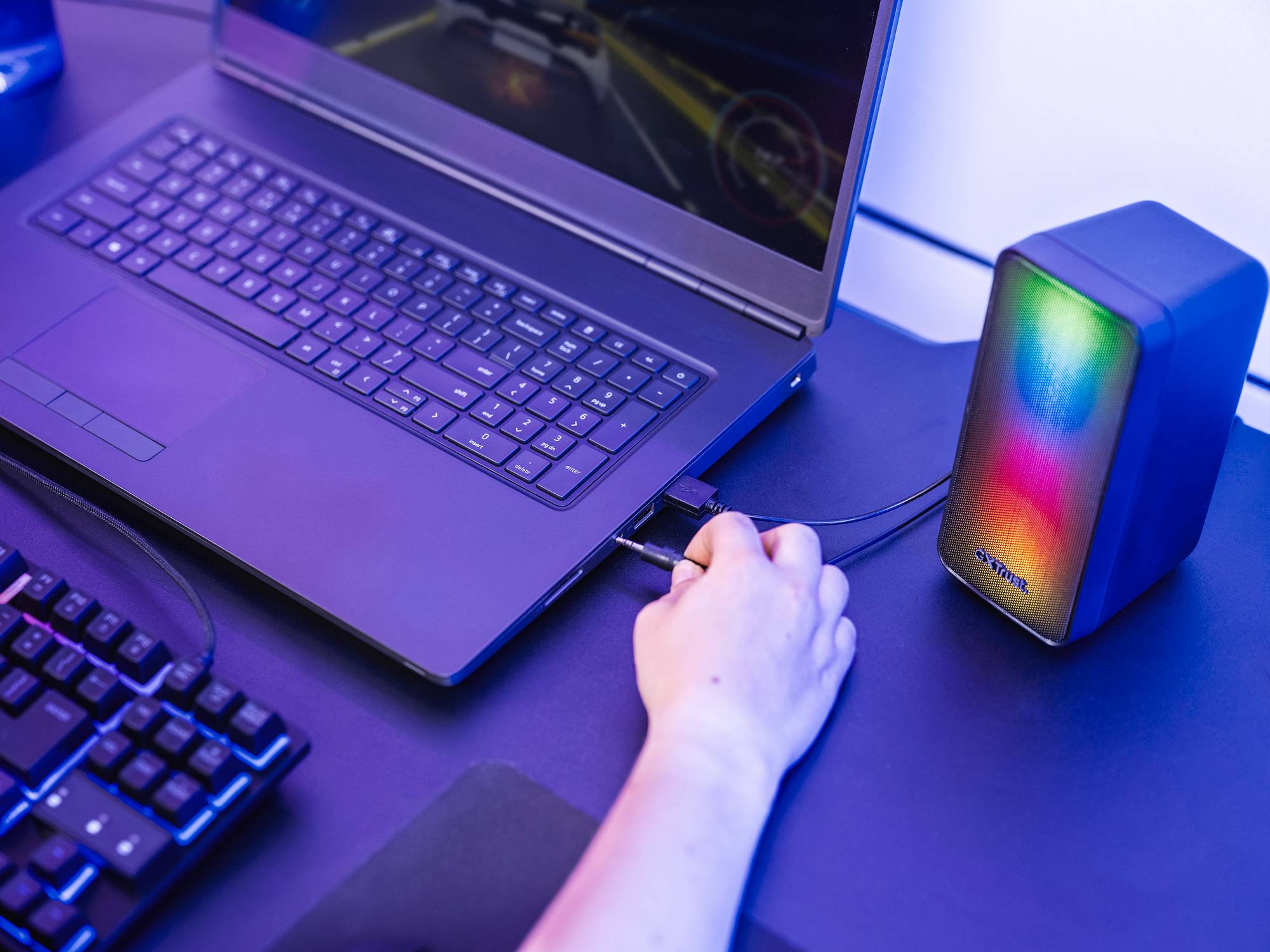 Otevřený laptop na stole, vedle něj svítící bluetooth reproduktor. Ruka zapojuje USB kabel do laptopu.