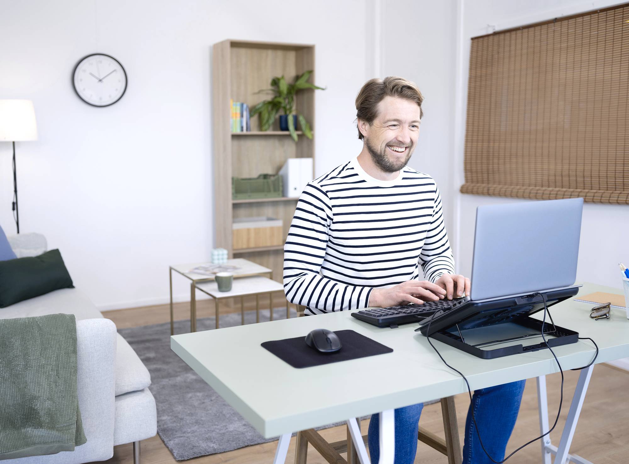 Muž v pruhovaném svetru se usmívá, zatímco pracuje na laptopu v moderní domácí pracovně s pohovkou a policí v pozadí.