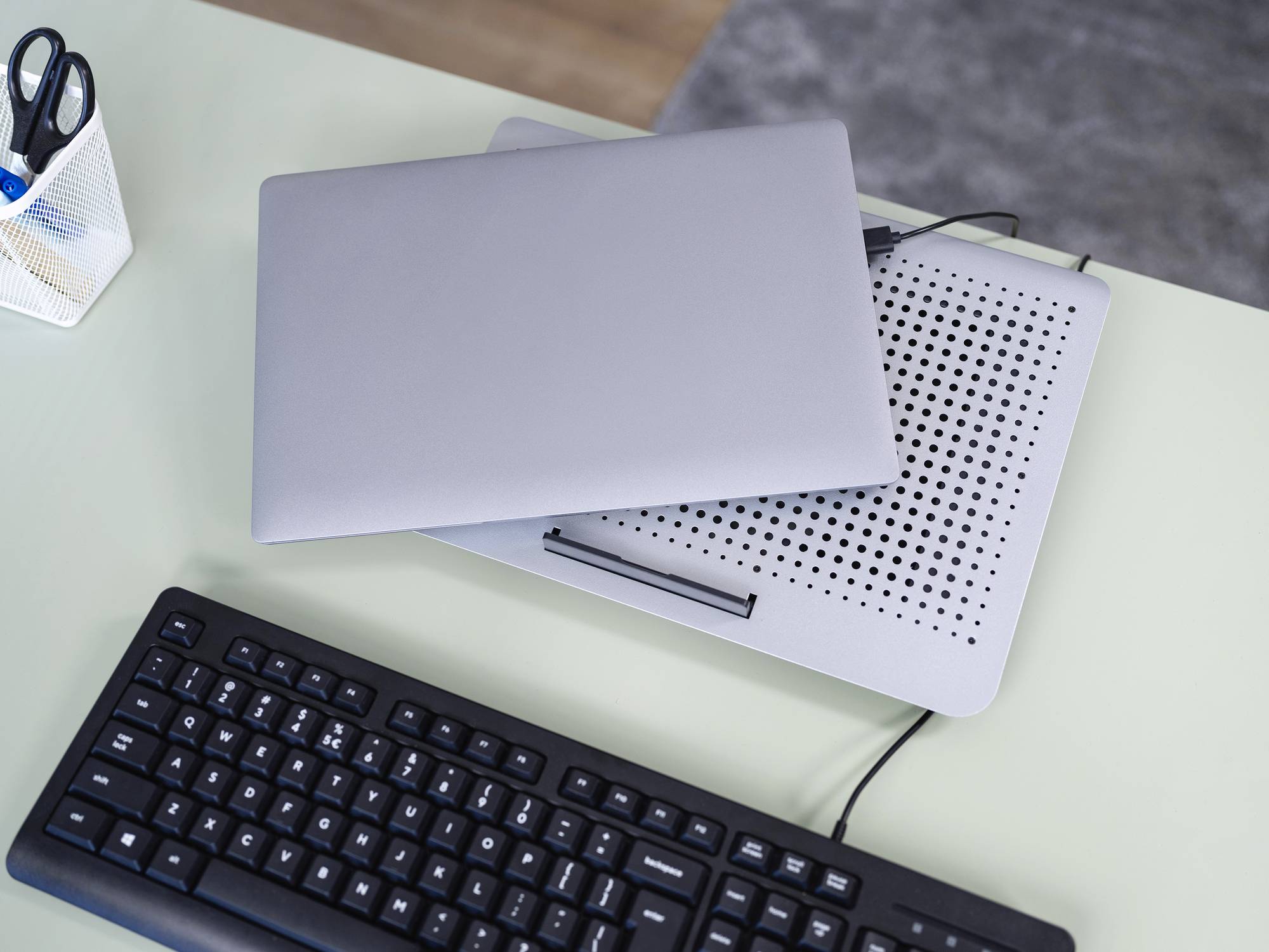 Uzavřený notebook na chladicí podložce na psacím stole vedle klávesnice. V pozadí stojí stojánek na tužky.