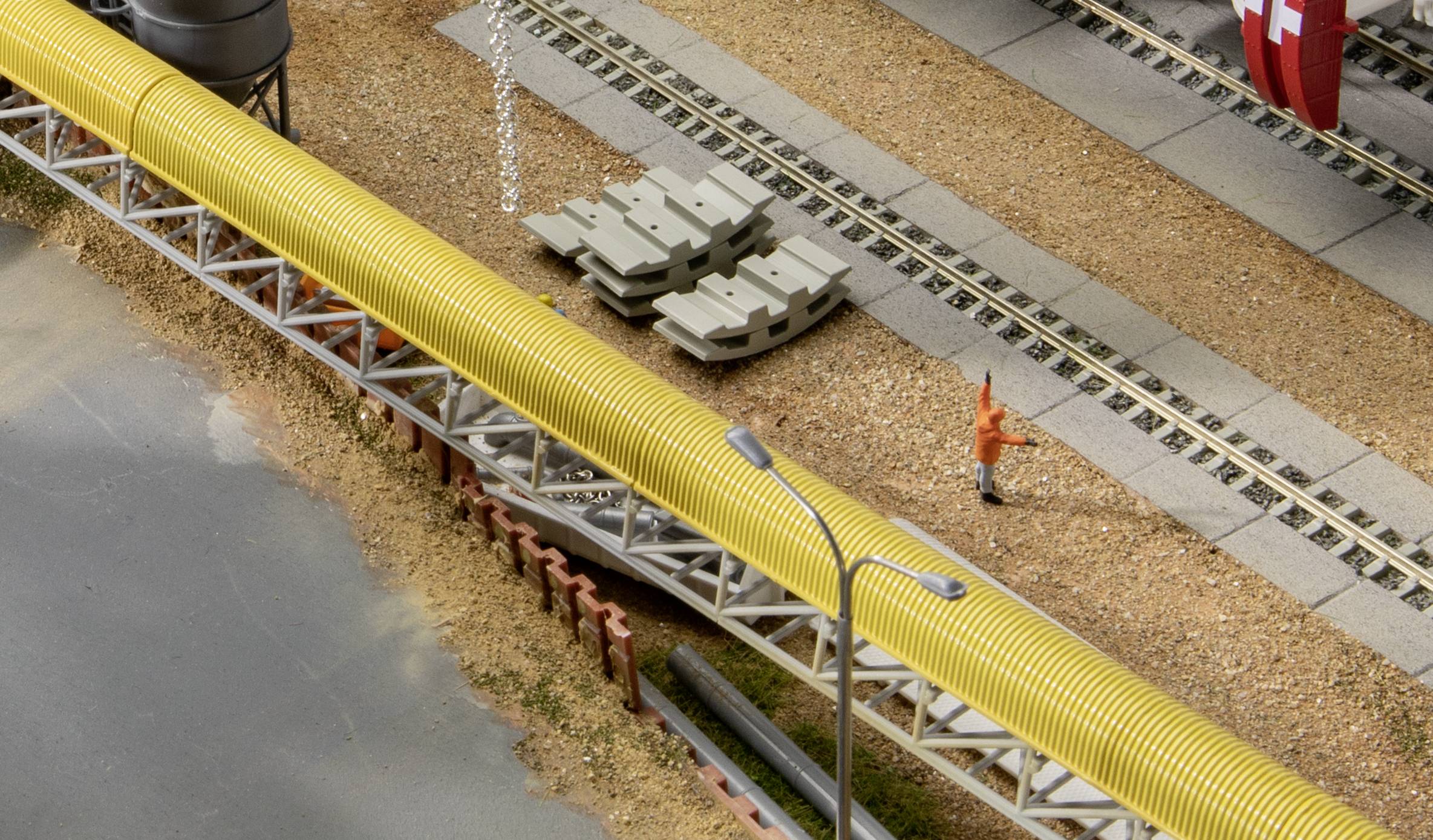 Miniaturní vlak jede po kolejích vedle žluté potrubní trasy. Dělník v oranžové bundě stojí poblíž.