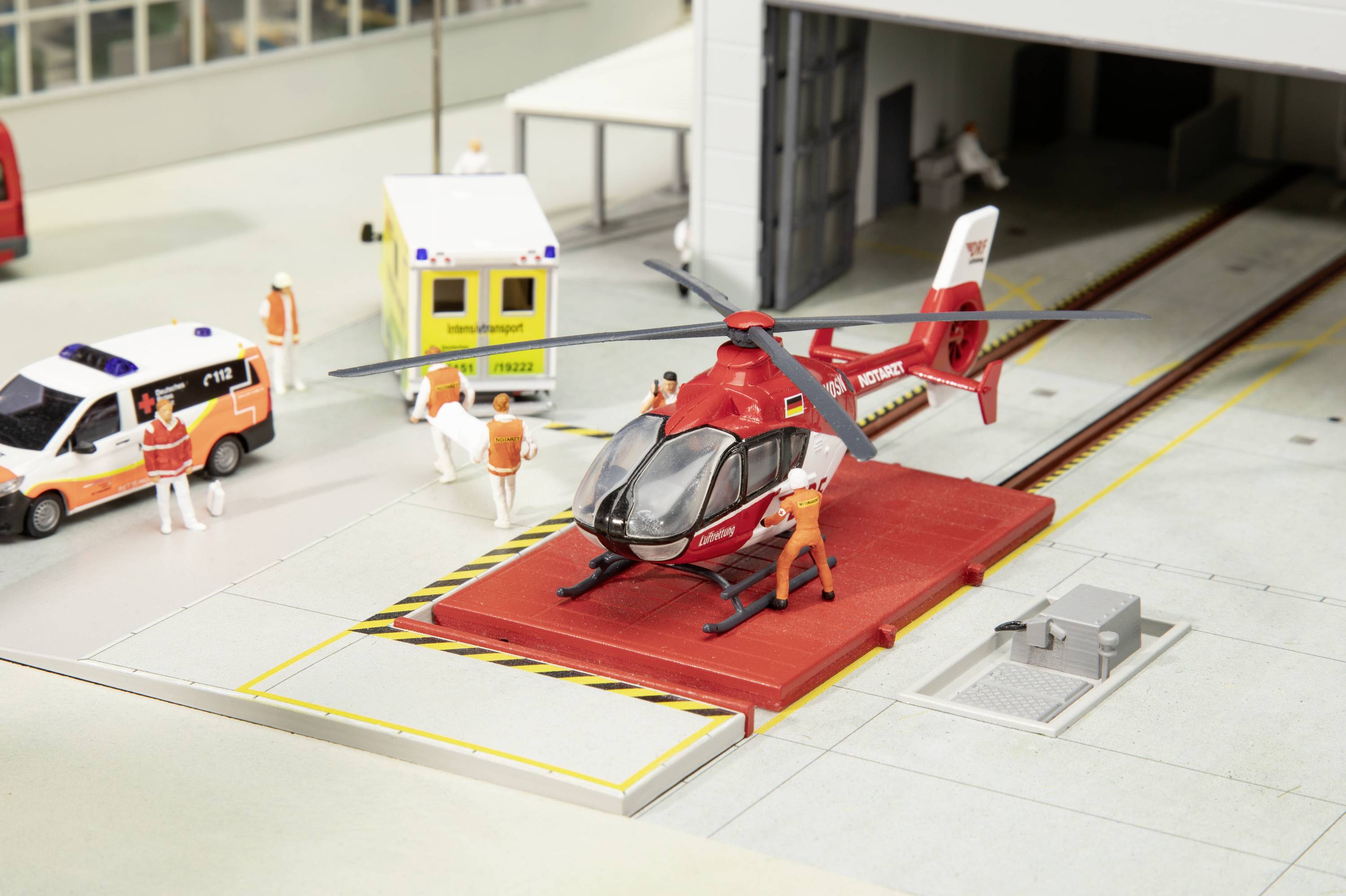 Model vrtulníku na přistávací plošině s figurkami v záchranářském oblečení, sanitkou a ambulancí v pozadí.