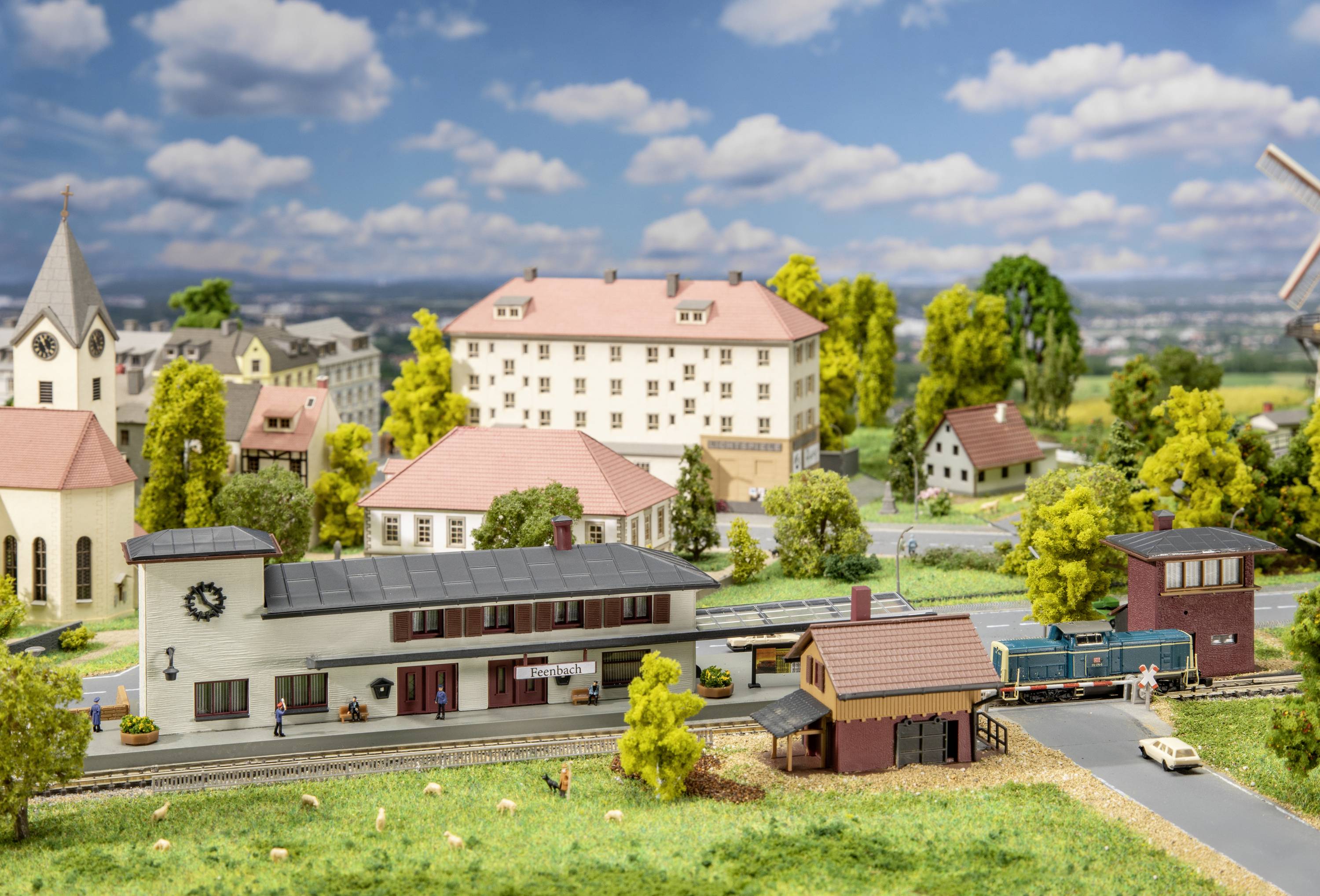 Modelová železniční krajina s nádražní budovou, malým vlakem a okolními budovami a stromy. Venkovské prostředí pod modrým nebem.