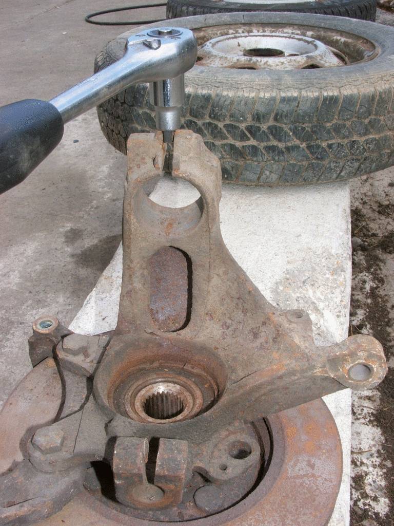 Detailní záběr ráčny pracující na zrezavělé součásti automobilu, s pneumatikou v pozadí.