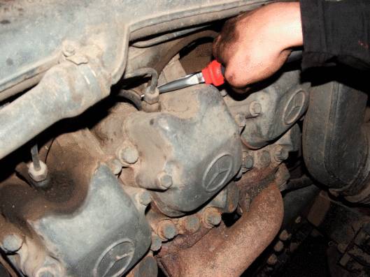 Mechanik kontroluje motor vozidla šroubovákem. Kryt motoru je prašný a opotřebovaný.