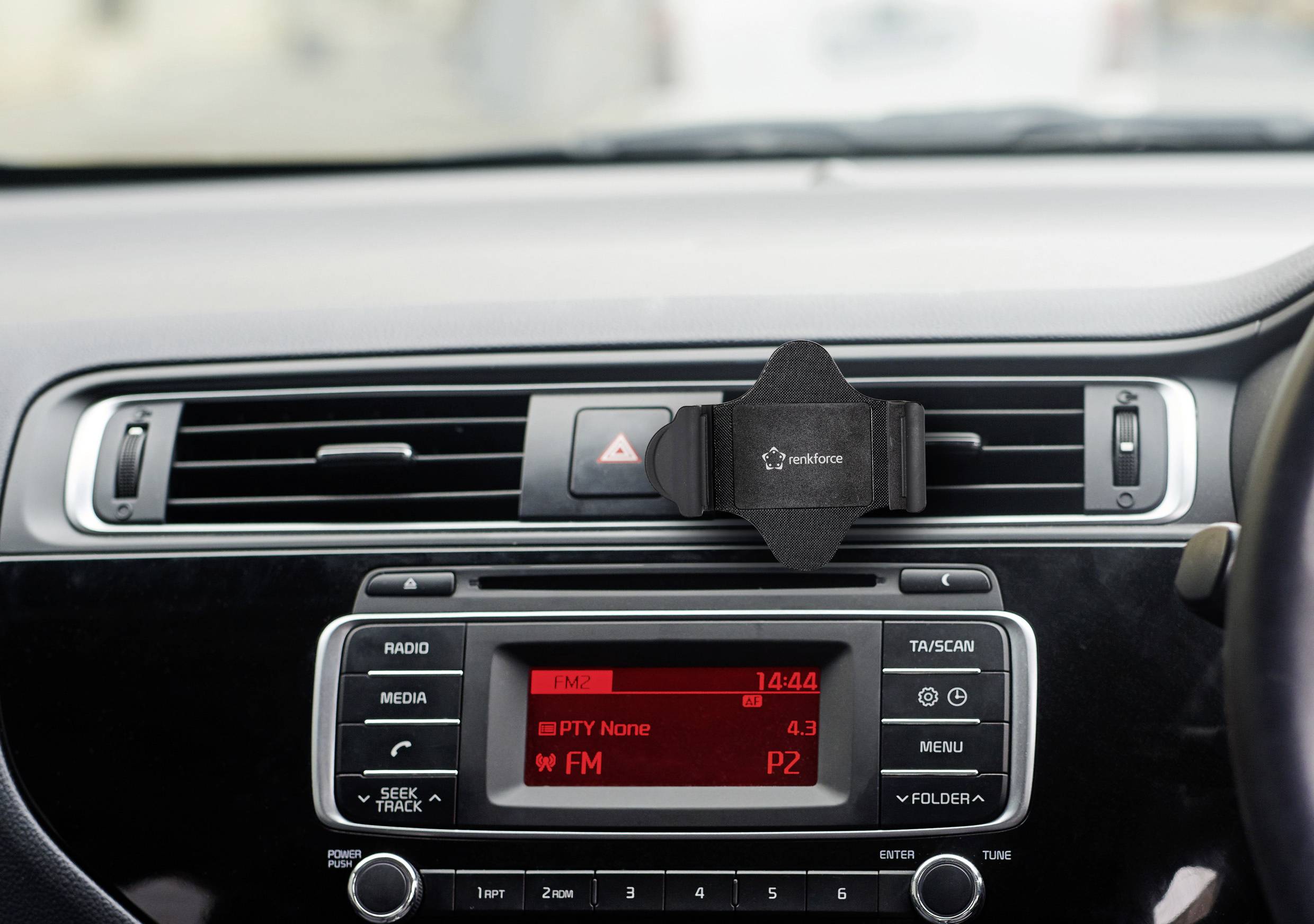 Palubní deska automobilu s rádiovým displejem. Zobrazuje čas '14:44', FM rádio zapnuté, stanice P2. Držák mobilního telefonu v ventilačních štěrbinách.