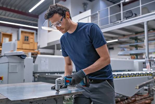 Pracovník v bezpečnostních brýlích a rukavicích řeže kovový plech ručním elektrickým nástrojem v průmyslovém workshopu.