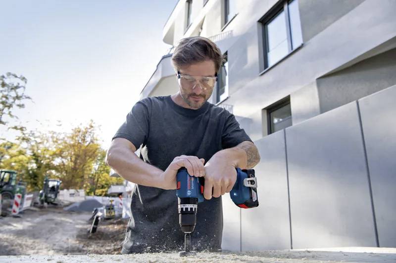 Dělník používající elektrickou vrtačku na staveništi, oblečený v ochranných brýlích, s budovou a stromy v pozadí.