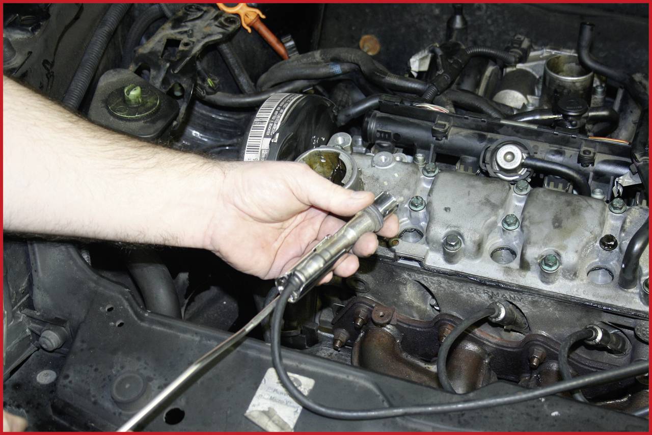Mechanik drží zapalovací svíčku před otevřeným blokem motoru, obklopen různými autodíly a nářadím.
