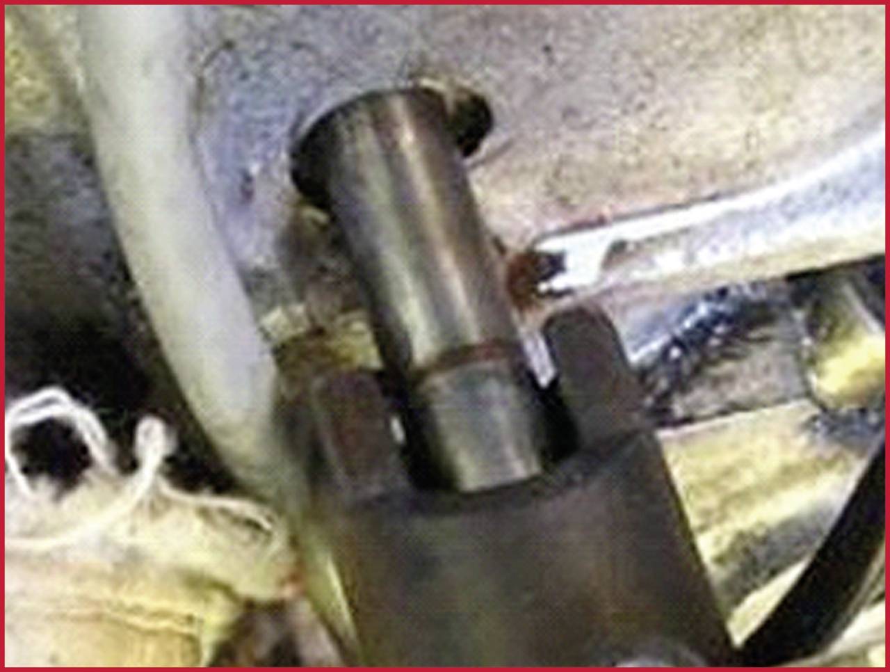 Kovový kolík nebo čep je zasazen do kovového držáku. Okolí zobrazuje hadice nebo kabely, pravděpodobně technické prostředí.