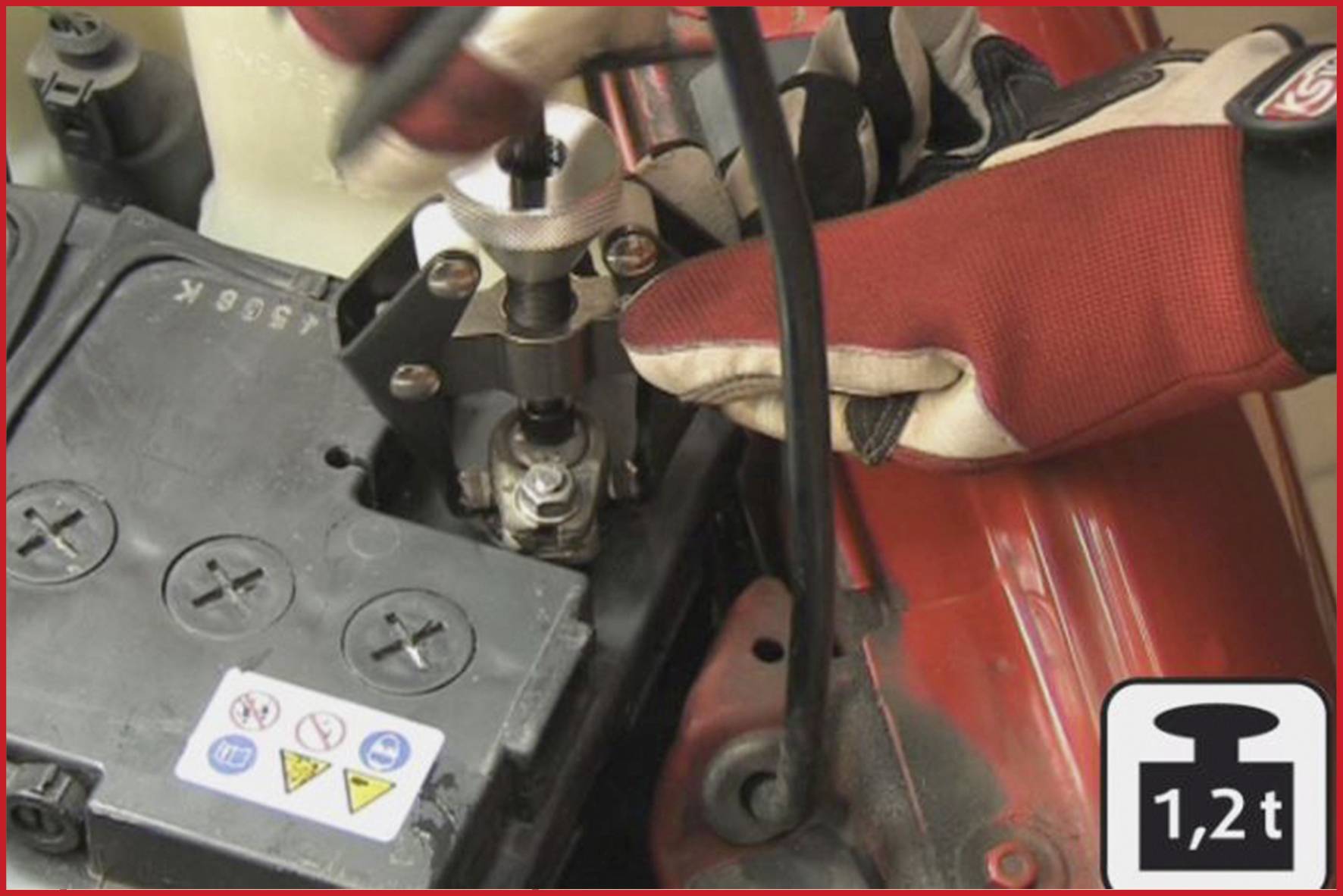 Červené svěrné kleště vytahují baterii z jejího držáku vozidla. Symbol zobrazuje '1,2t', což indikuje nosnost.