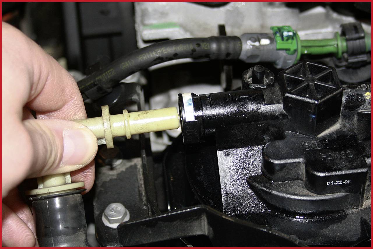 Jedna ruka drží žlutou plastovou hadici a zasunuje ji do černé mechanické součástky v motorovém prostoru.