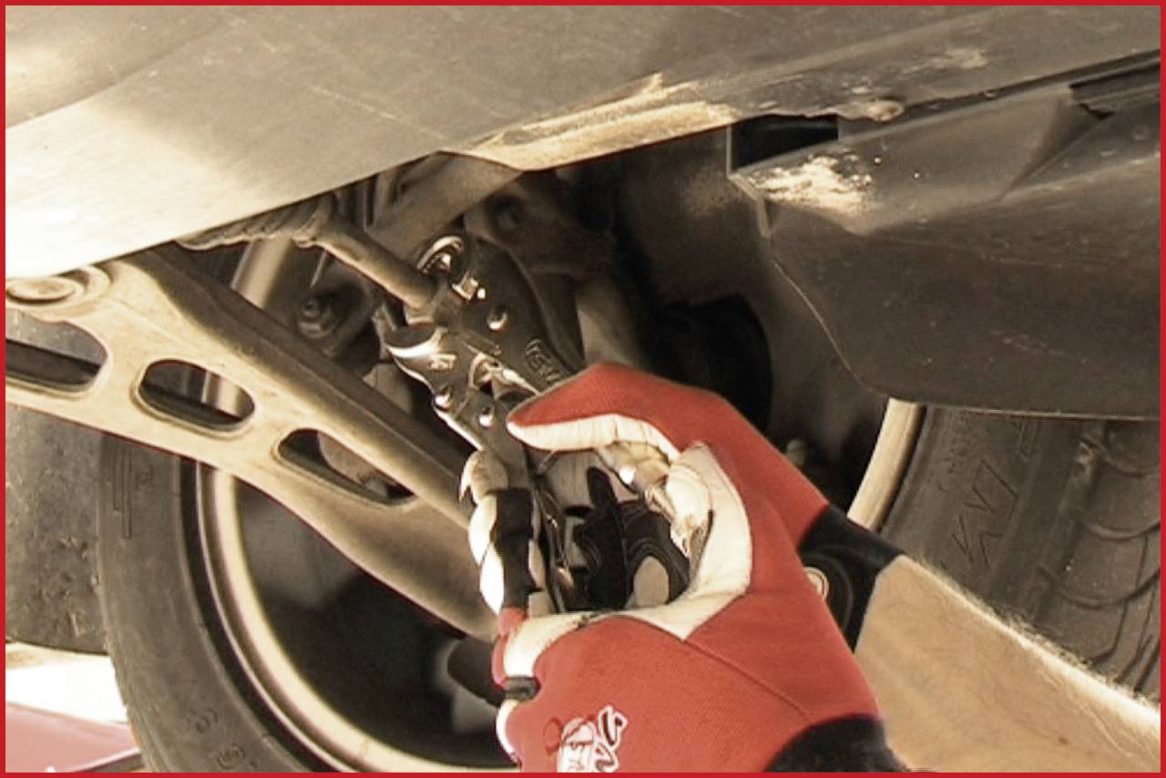 Osoba v červených rukavicích používá kleště, aby pracovala na části vozidla pod zdviženým autem.