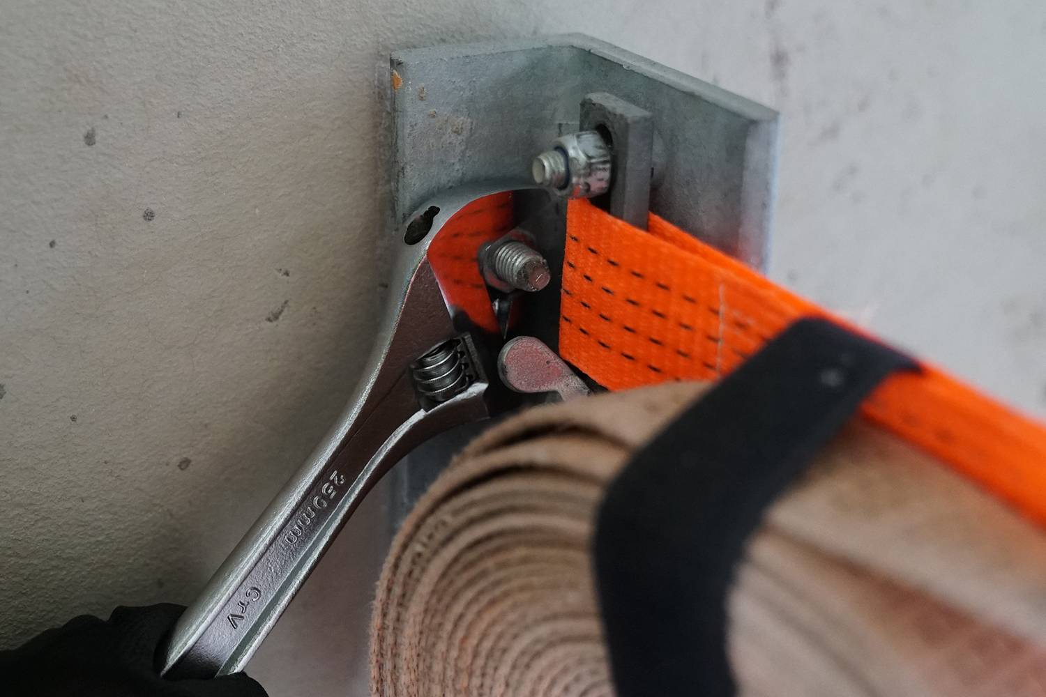 Francouzský klíč utahuje šroub, aby upevnil oranžový upínací popruh na kovovém držáku. Vlevo je viditelná rukavicí chráněná ruka.