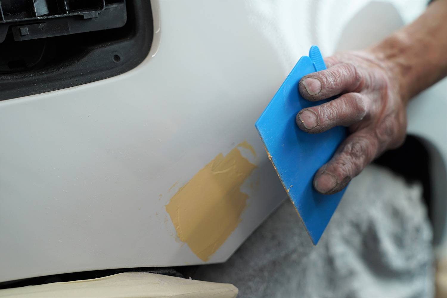 Jedna ruka nanáší žlutý tmel na bílou karoserii automobilu. Tmel je roztírán modrým plastovým nástrojem.