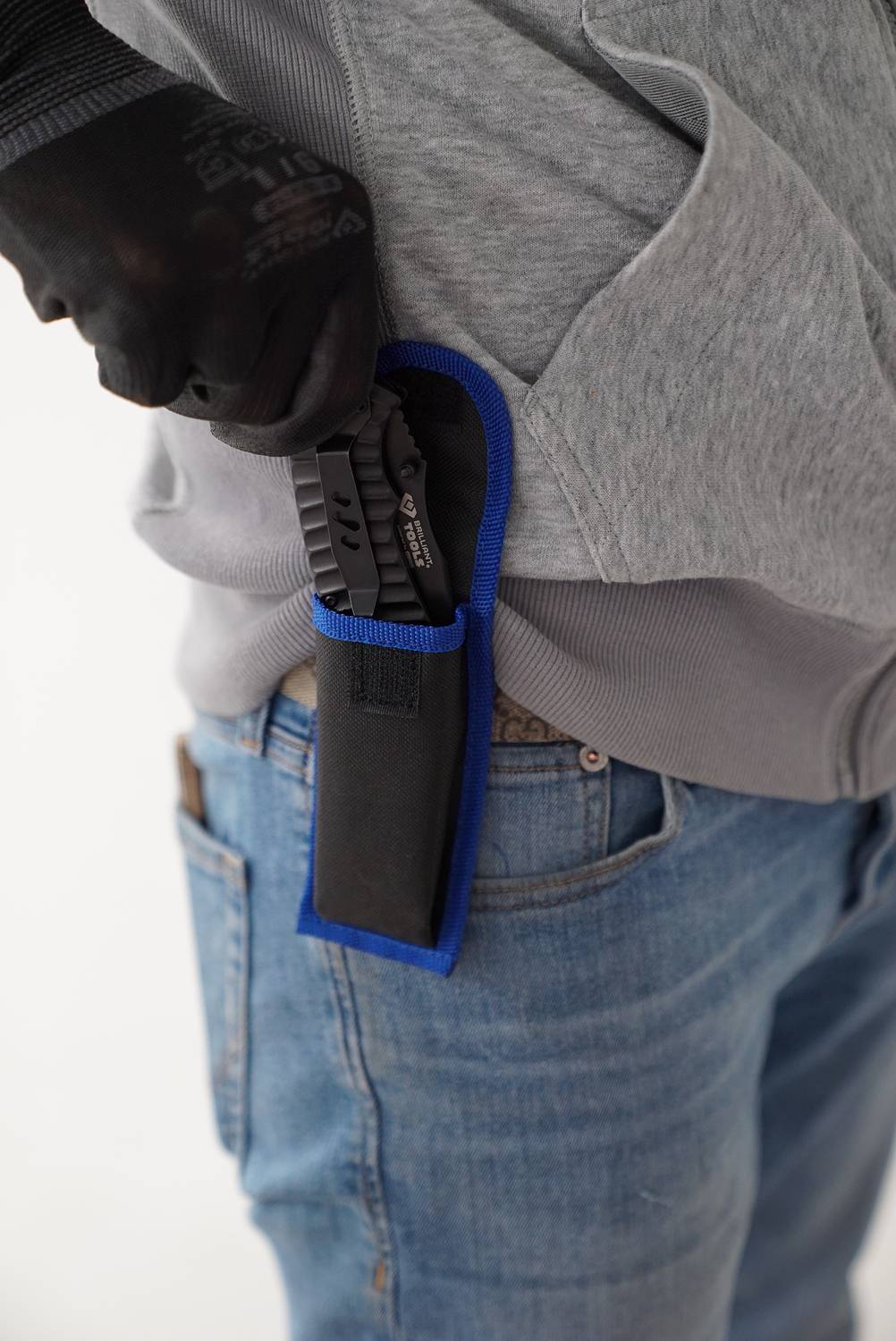 Osoba v modrých džínách má na sobě šedou mikinu a opatrně vytahuje černý nůž z modrého pouzdra připevněného k opasku.