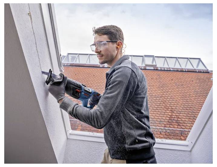 Muž v ochranných brýlích a rukavicích používá elektrický nástroj k řezání zdi na střeše, přičemž v pozadí jsou viditelné střechy s oranžovými taškami.