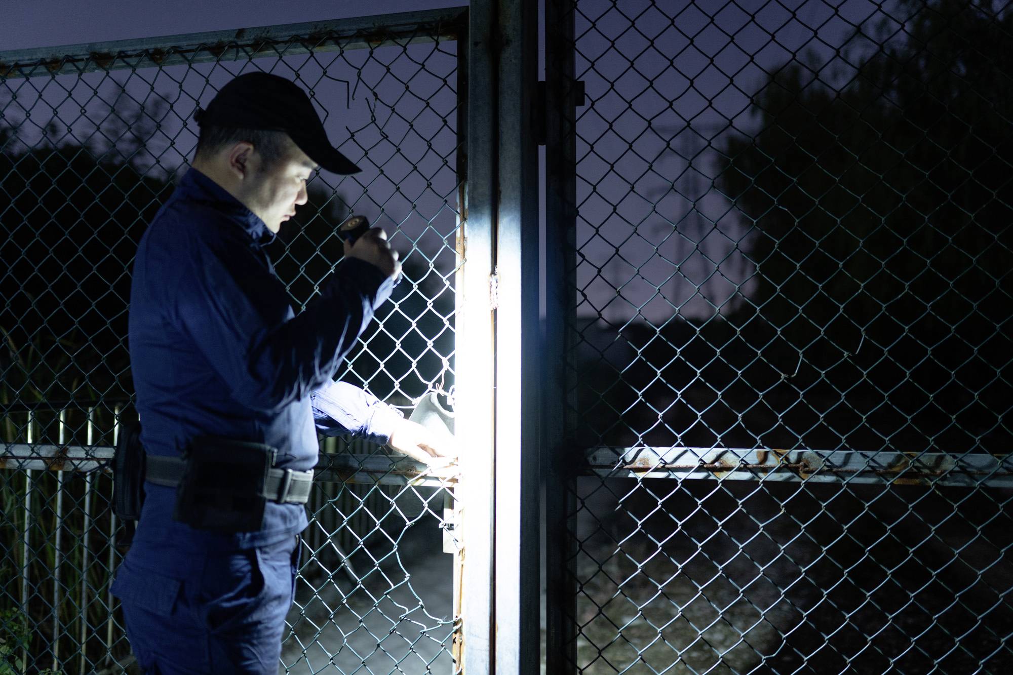 Bezpečnostní pracovník v noci prozkoumává uzamčenou mříž pomocí kapesní svítilny.