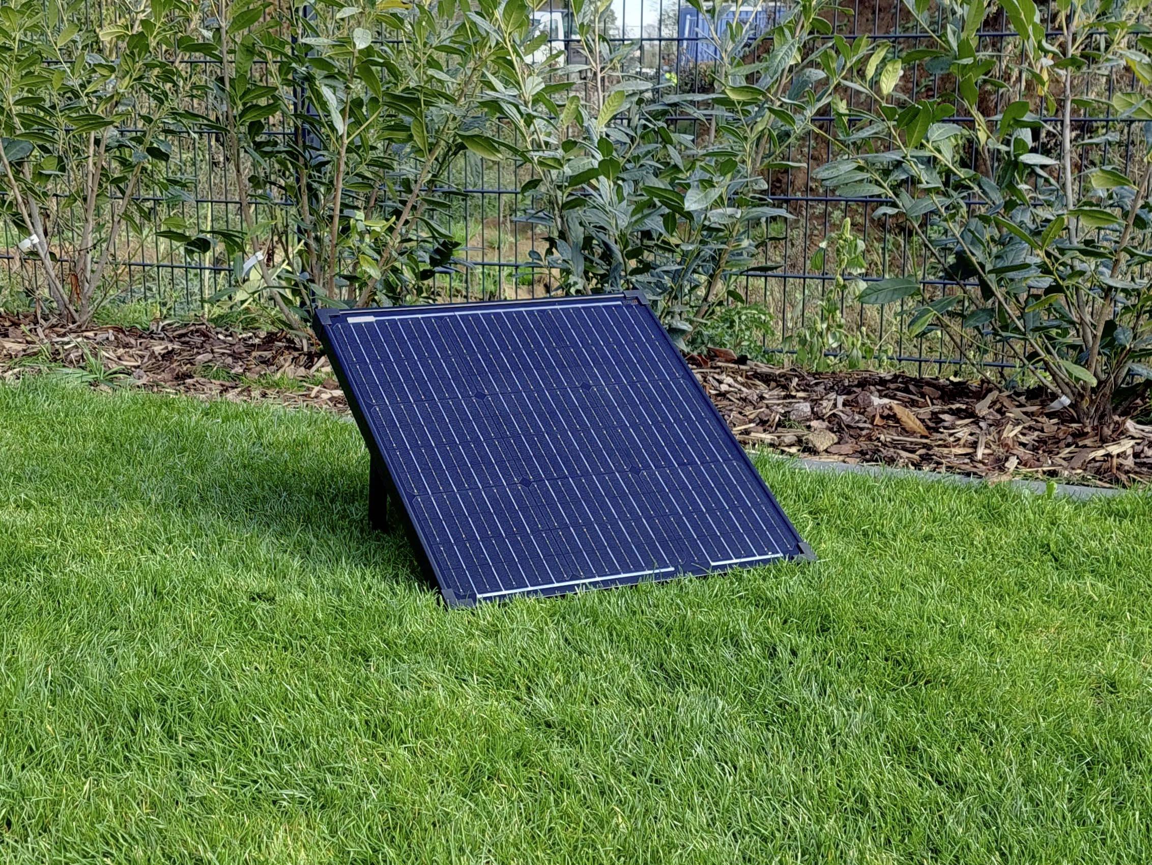Solární panel stojí šikmo na zelené trávě v zahradě. V pozadí jsou vidět rostliny a plot.