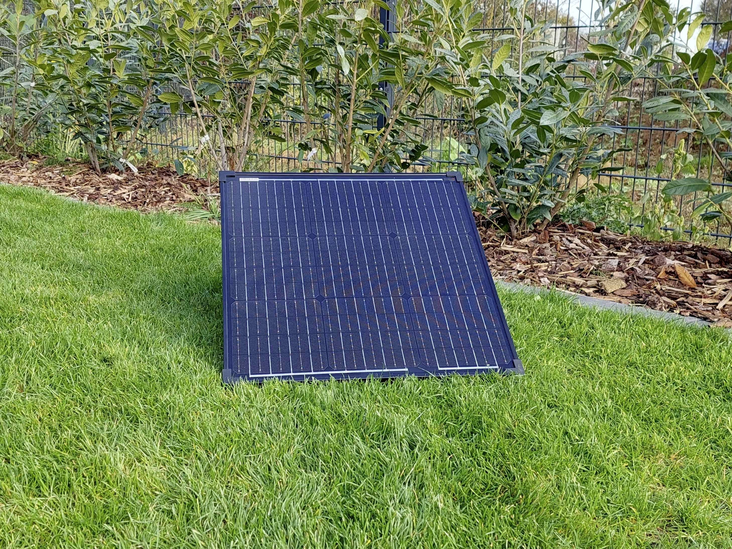 Solární panel stojí vzpřímeně na travnaté ploše, obklopen nízkými keři.