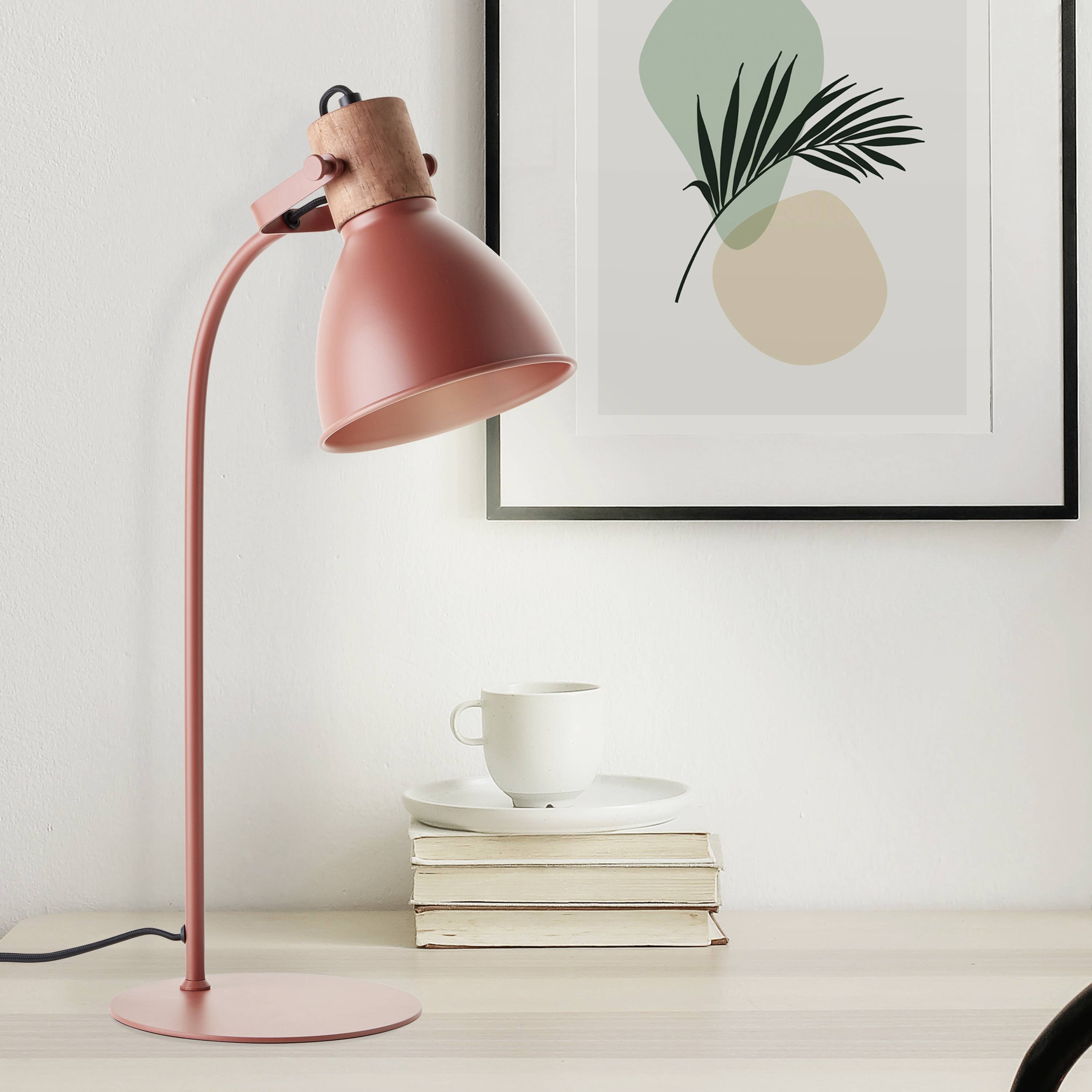 Růžová stolní lampa stojí na stole vedle stohu knih, bílého hrnku a abstraktního obrazu v pozadí.
