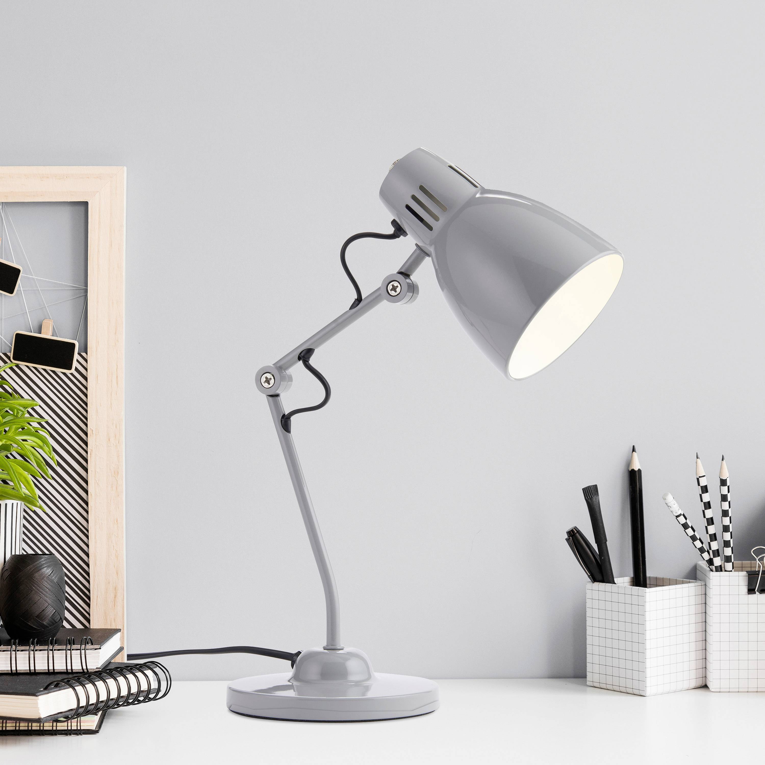 Stolní lampa na psacím stole osvětluje notes a stojánek na tužky. V pozadí rám s rostlinou.