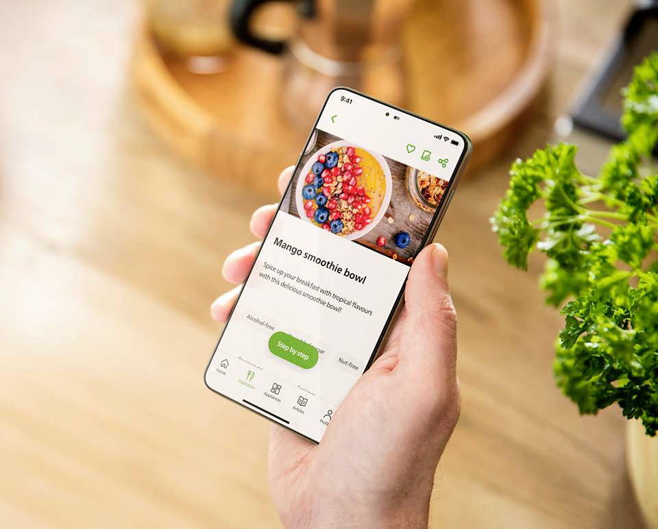 Jedna ruka drží smartphone s aplikací zobrazující recept na 'Mangovou smoothie mísu'. V pozadí je vidět petržel.
