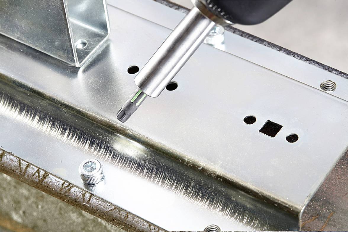 Šroubovák utahuje šroub do kovového dílu. Je vidět lesklý kovový plech a částečně zašroubovaný šroub.