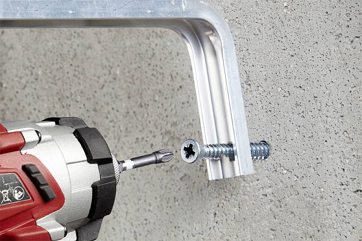 Akumulátorový šroubovák zašroubovává šroub přes kovový úhelník do betonové stěny. Vrták otáčí šroubem.