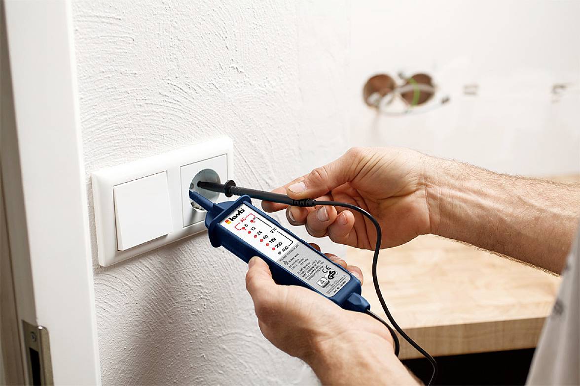 Osoba testuje elektrickou zásuvku napěťovým testerem, který ukazuje, zda obvodem prochází elektrický proud. V pozadí jsou viditelné holé elektrické vodiče.