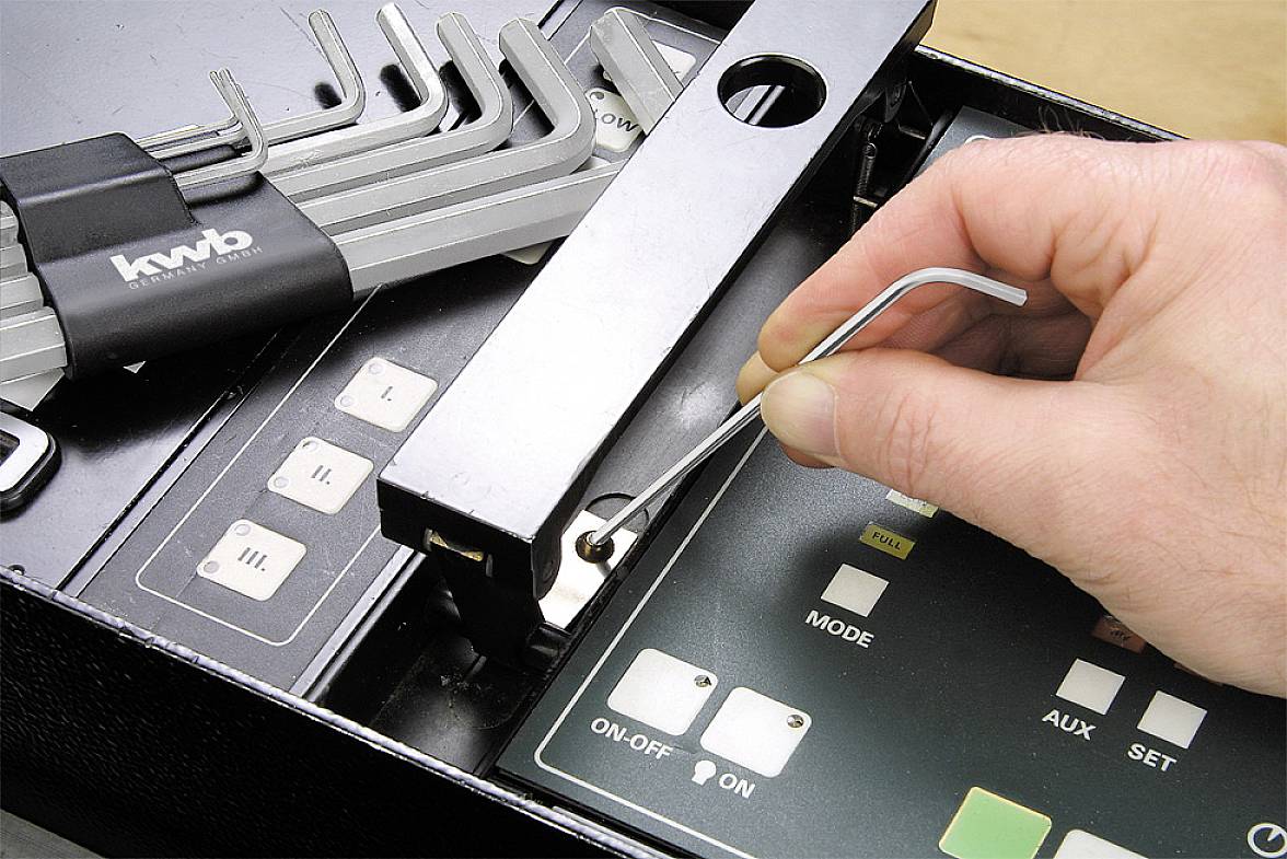 Jedna ruka používá inbusový klíč k otevření zámku. Je vidět také montážní kufřík s dalšími inbusovými klíči a ovládací rozhraní.