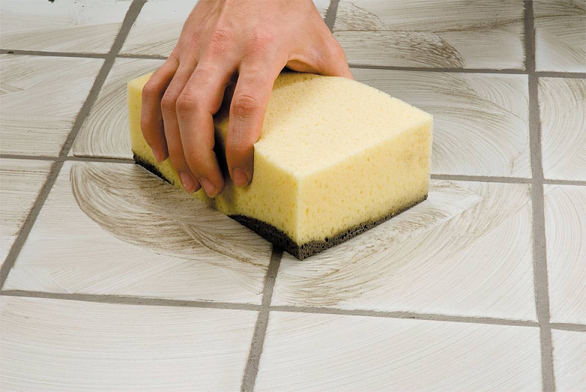 Ruka čistí špinavé dlaždice na podlaze žlutou houbou.