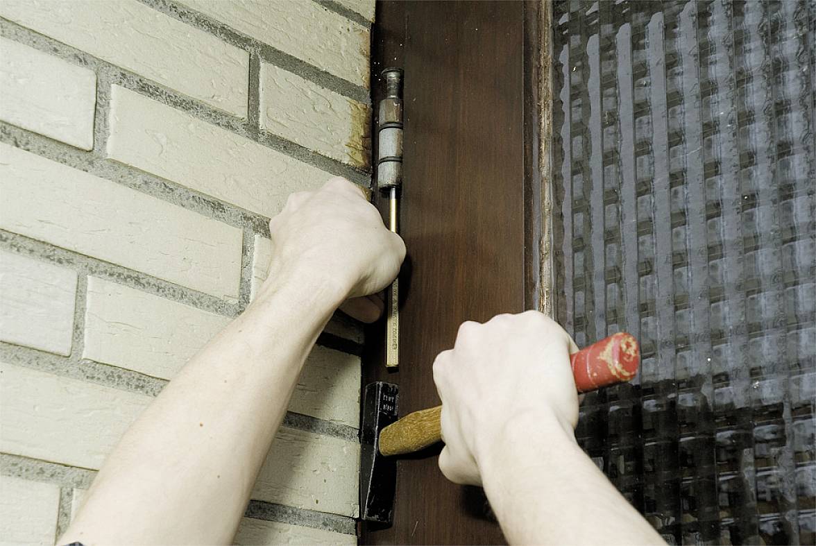 Dvě ruce používají kladivo a sekáč k opracování pantů dveří nebo okna na červenohnědém rámu.